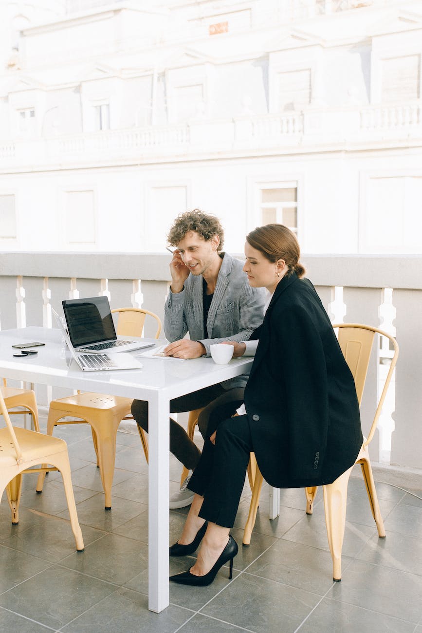 Kobieta i mężczyzna wspólnie pracują przy komputerach siedząc przy stoiku na patio budynku.